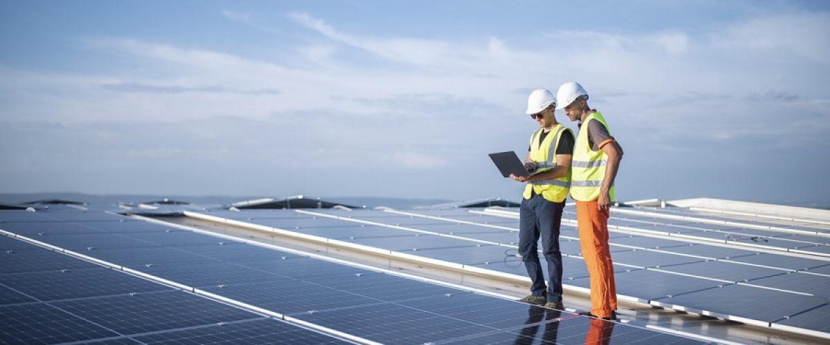 Team of engineers using technology while installing solar panels on a roof of warehouse.